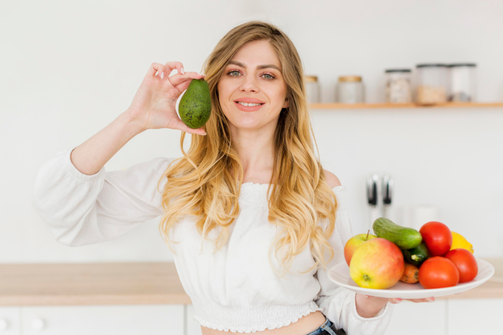 Smiling woman holding an avocado and a plate of fresh fruits and vegetables, showcasing a healthy hair diet for vibrant hair.