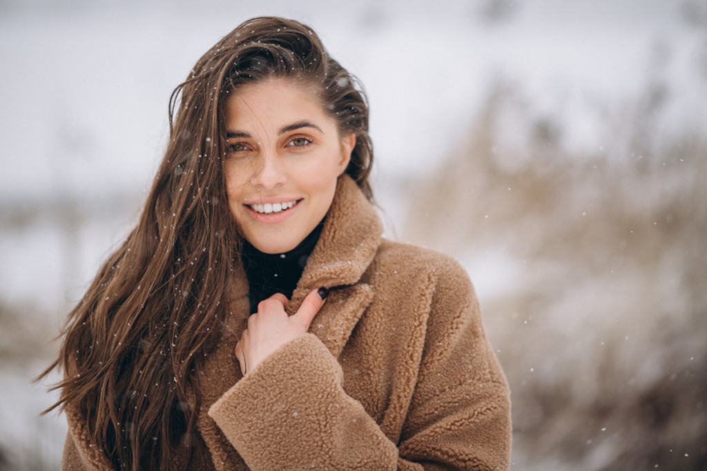 Smiling woman with long brown hair in a warm coat during winter, showcasing winter hair care in a snowy outdoor setting.