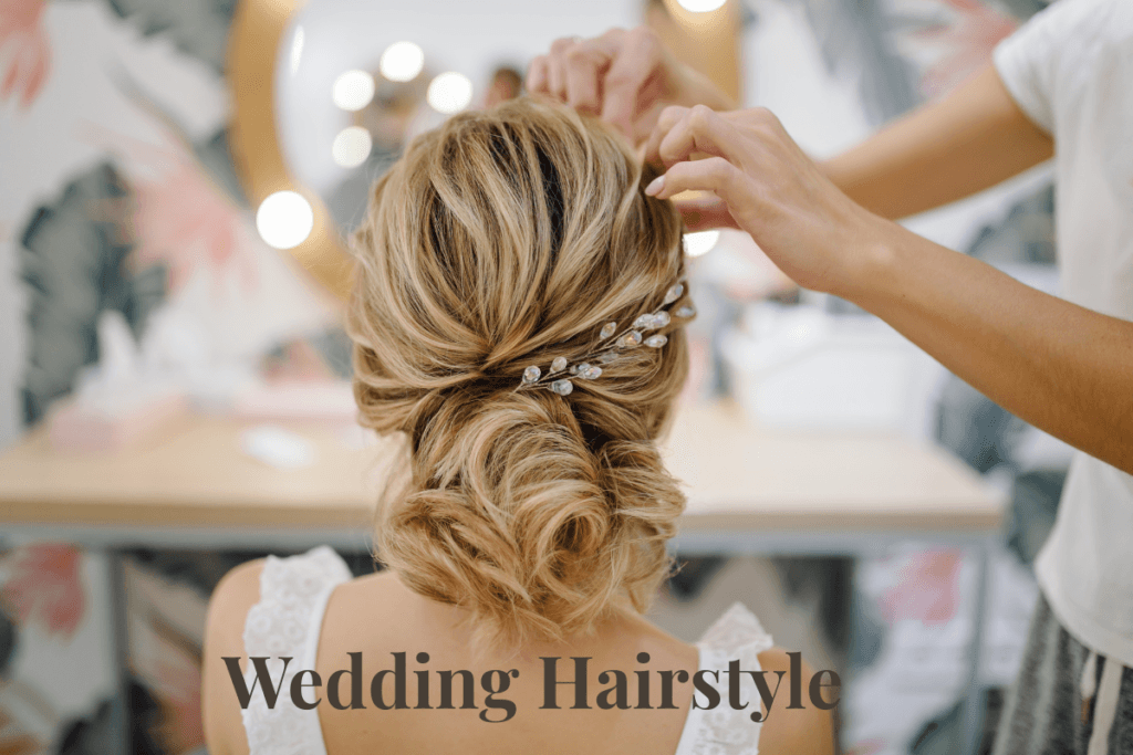 Stylist arranging elegant low bun wedding hairstyle with jeweled hairpin on bride’s blonde hair.