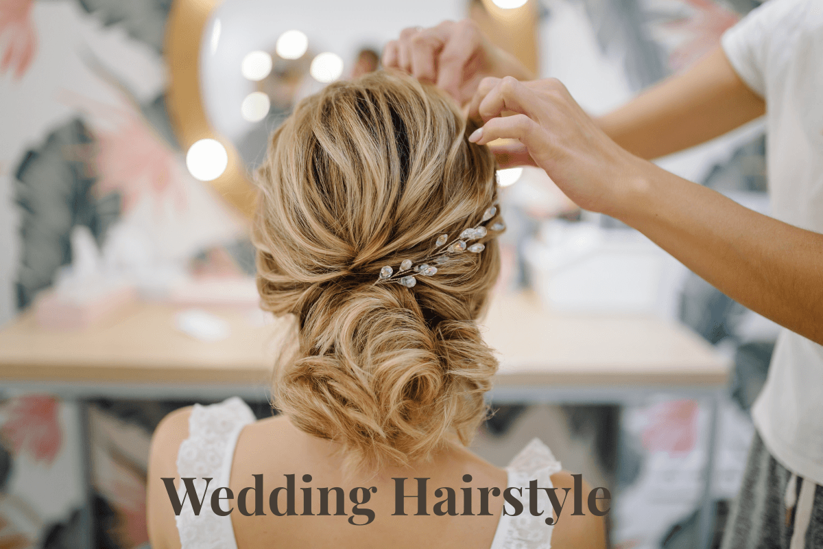 Stylist arranging elegant low bun wedding hairstyle with jeweled hairpin on bride’s blonde hair.